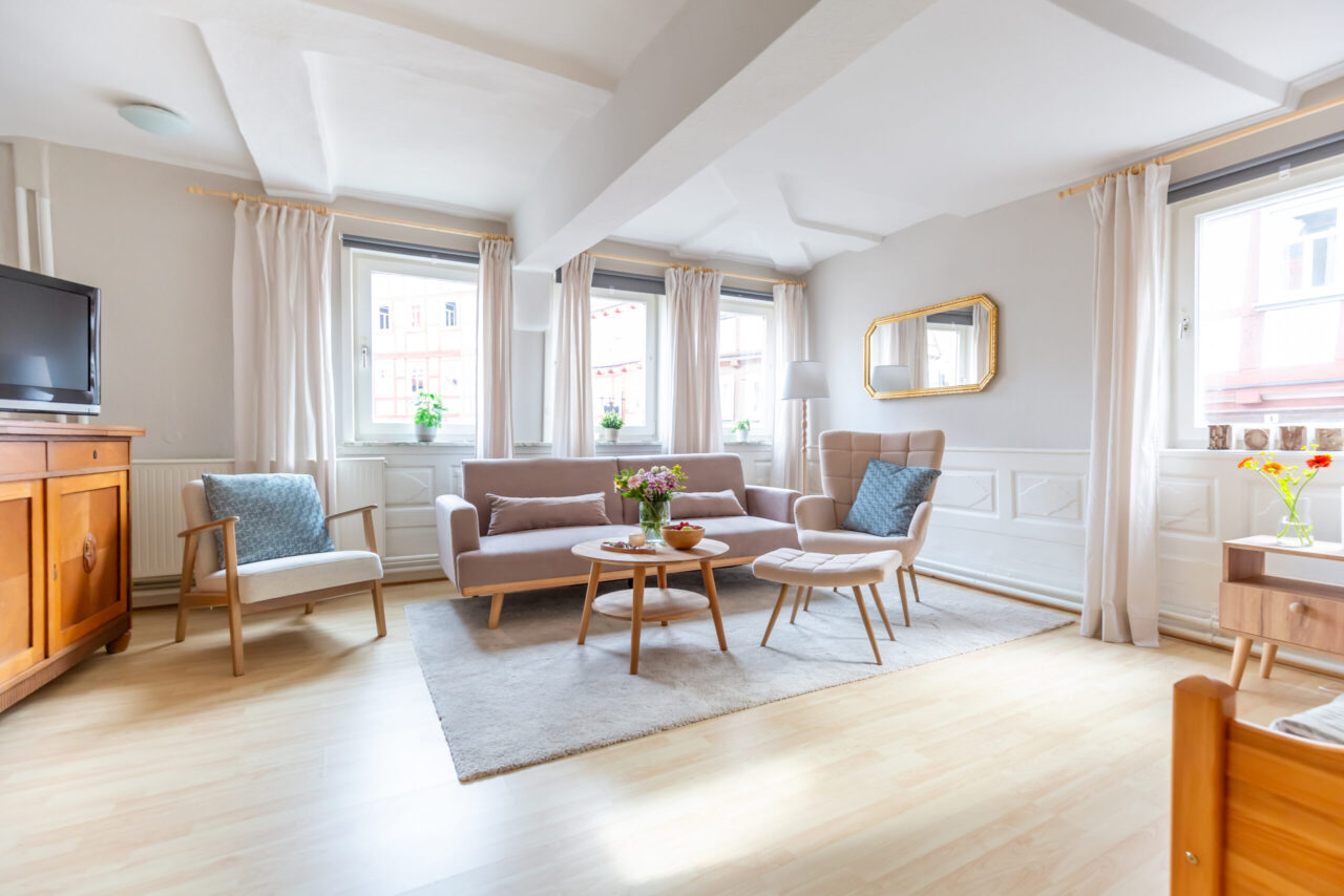 Lichtdurchflutetes Wohnzimmer einer Ferienwohnung: Sofaecke mit Ohrensessel und Hocker, Holzcouchtische, Spiegel, helle Vorhänge und Parkett.