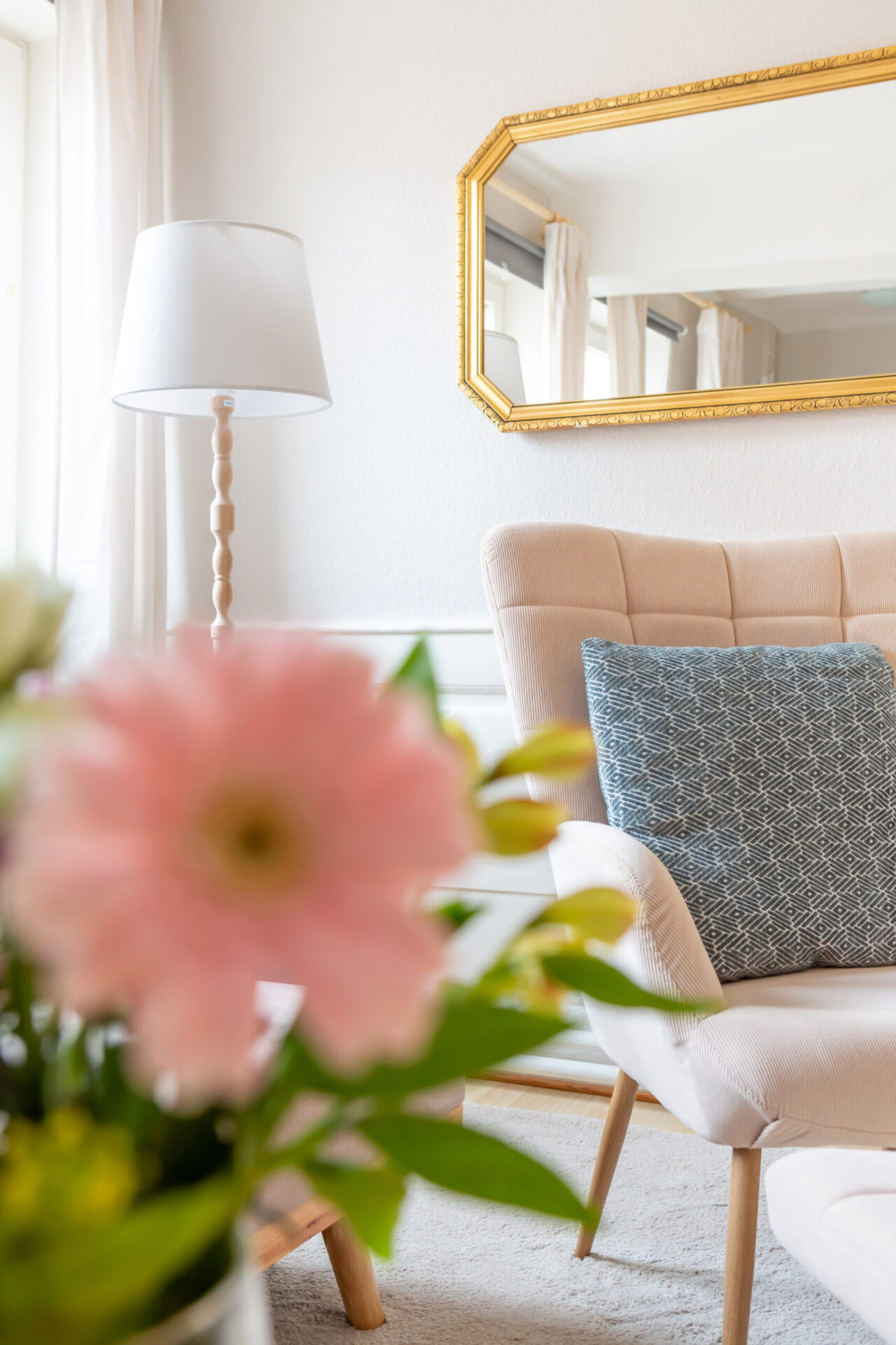 Helles Wohnzimmer-Detail einer Ferienwohnung: bequemer beiger Sessel mit blauem Kissen, goldener Wandspiegel, helle Stehlampe und frische Blumen im Vordergrund.