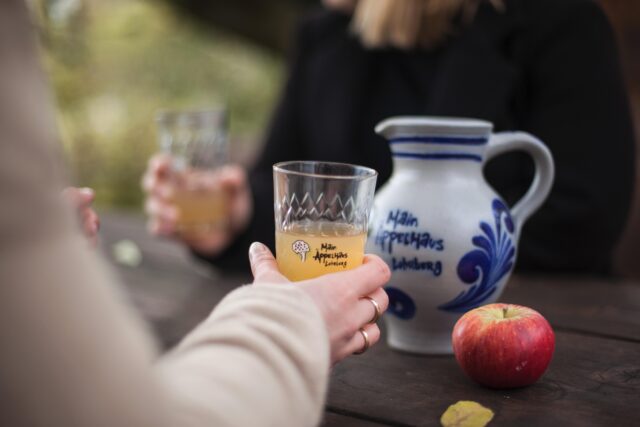 Apfelwein im Gerippten, gehalten von zwei Personen; grauer-blauer Bembel und roter Apfel auf Holztisch.