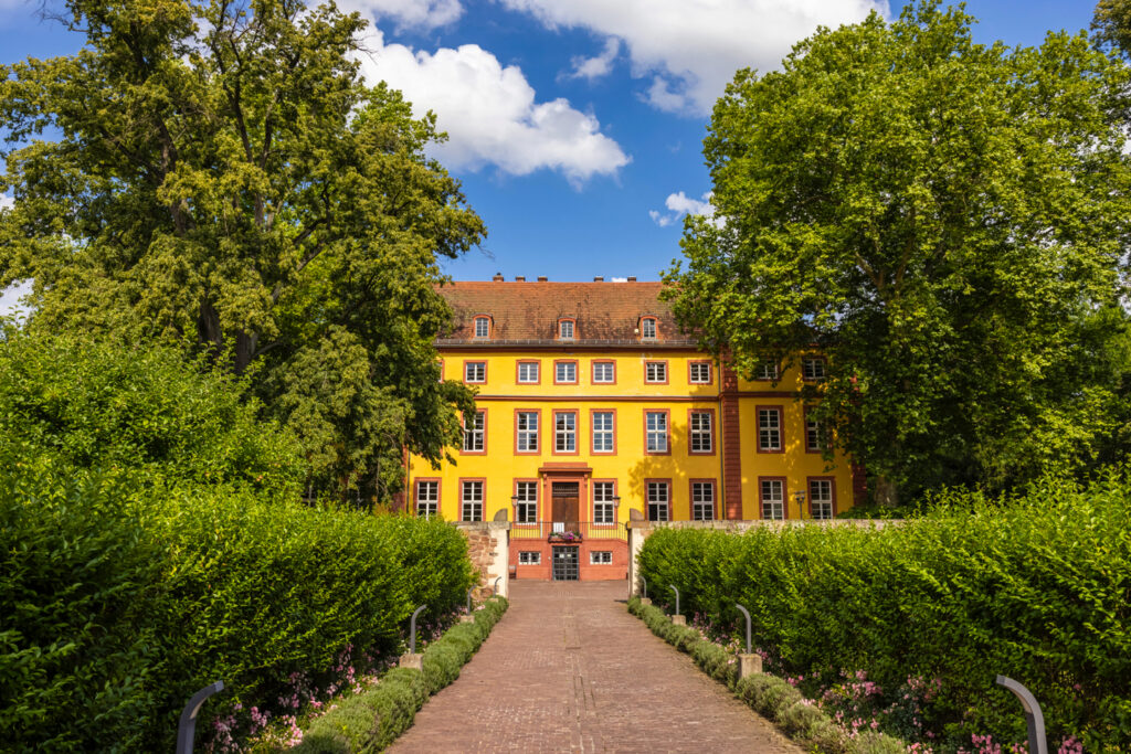 Von zwei großen Bäumen auf beiden Seiten gesäumt und mit blauem Himmel im Hintergrund, erhebt sich ein mehrstöckiges, leuchtend gelbes Barockes Gebäude.