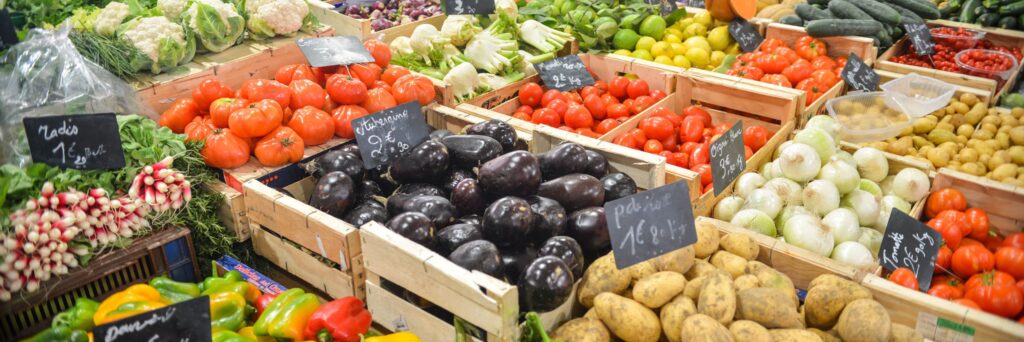 Wochenmarktstand mit Holzsteigen voller buntem Gemüse – Tomaten, Auberginen, Radieschen, Paprika, Zwiebeln und Kartoffeln – dazu kleine Tafeln mit Euro-Preisen.