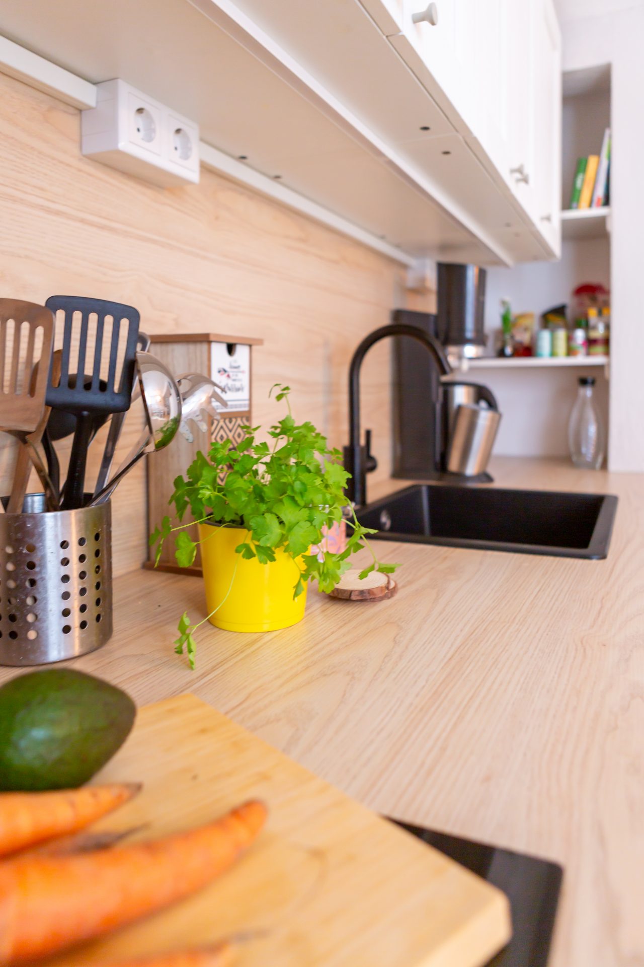 "Detailansicht der hellen Küchenzeile mit Spüle, schwarzer Armatur, Küchenutensilien und frischen Kräutern