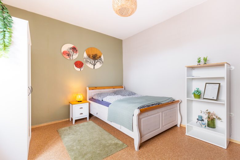 Gemütliches Gästezimmer zur Vermietung mit hellem Bett, Nachttisch, Regal und dekorativer Wandgestaltung.